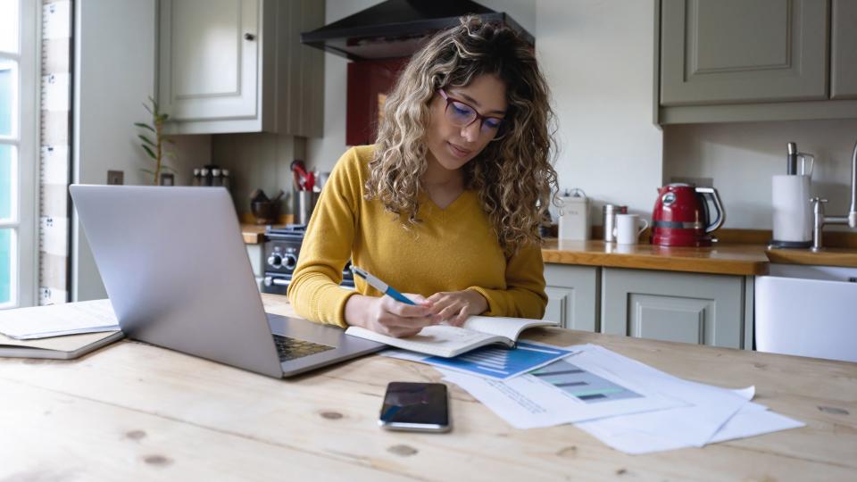 Beautiful young woman with curly hair working from home writing on notepad and using laptop and documents - Lifestyles.