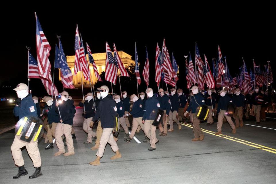 Grupo de hombres vestidos con camisas azules, pantalones caqui y pasamontañas que portan la bandera estadounidense