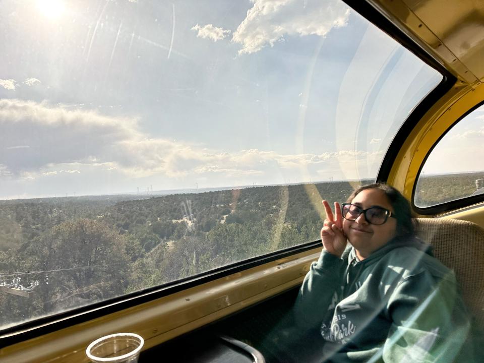 Athena, wearing a green sweatshirt and sunglasses, poses next to the large 360-degree observation window on the Grand Canyon Railway's luxury-dome car.