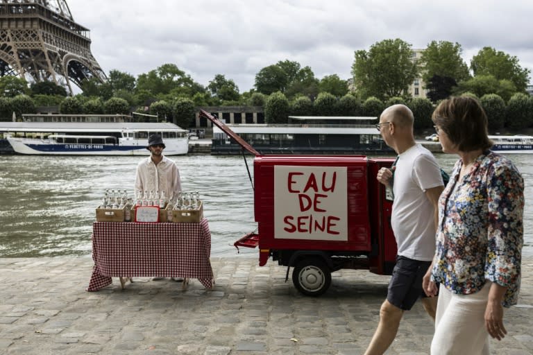 Dos transeúntes pasan frente a la instalación del artista callejero James Colomina de venta de agua del Sena embotellada, el 10 de julio de 2024 junto al río de París, a la altura de la Torre Eiffel (Olympia de Maismont)