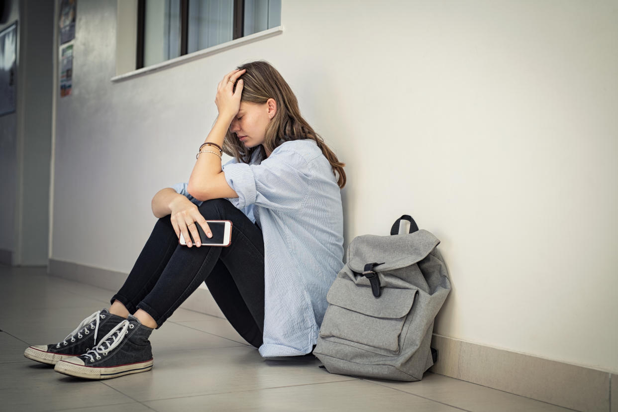 A high school girl sits in a school hallway looking forlorn, with hand to head and phone in hand
