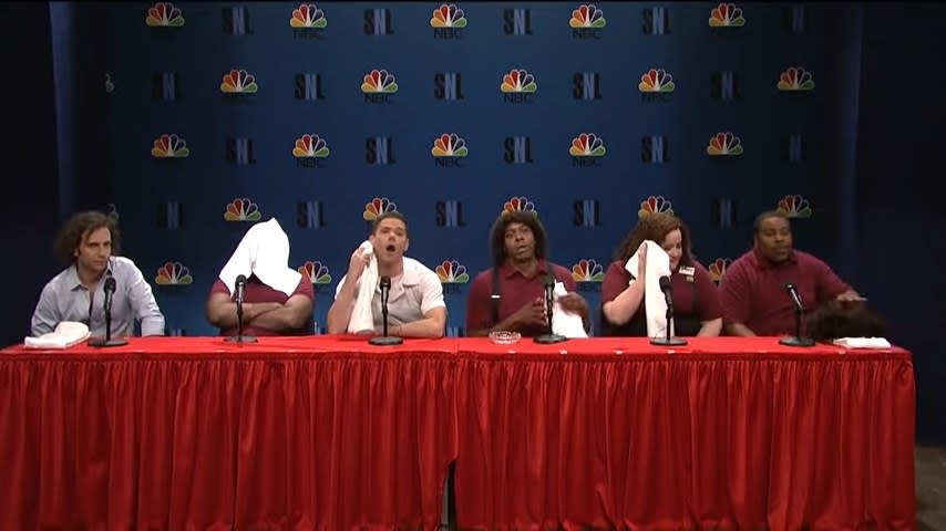 Dave Chappelle, Kenan Thompson, Aidy Bryant, Mikey Day, Leslie Jones, and Kyle Mooney sitting at a table with towels and microphones in "Saturday Night Live"