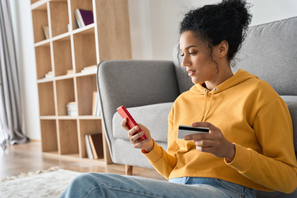 Young adult African American female consumer holding credit card and smartphone sitting on floor at home doing online banking transaction. E commerce virtual shopping, secure mobile banking concept.