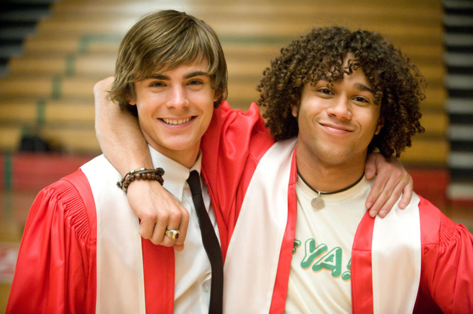Zac Efron and Corbin Bleu smiling with arms around each other, wearing graduation robes in a gymnasium