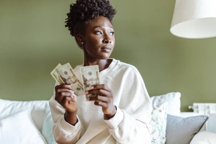 Woman in a white sweater holding cash, looking pensively to the side. Lamp and pillows in the background. Names not provided