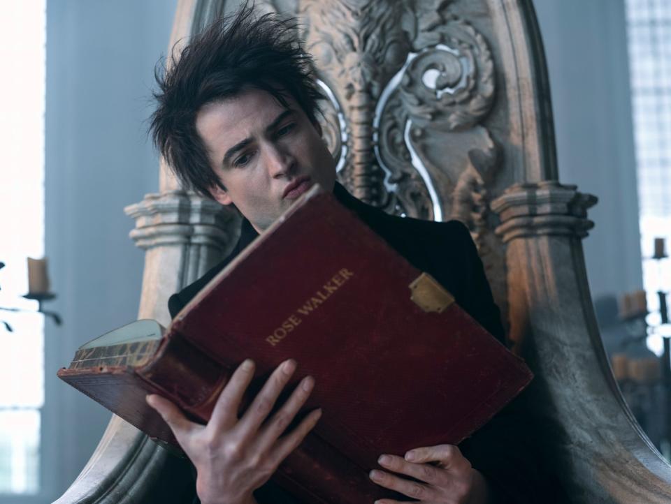 tom sturridge as dream in the sandman, a young man with wild brunette hair, sitting on a stone throne and holding a large, red tome with the text "rose walker" on the front. his hands are splayed across the back of the book and he is reading it intently
