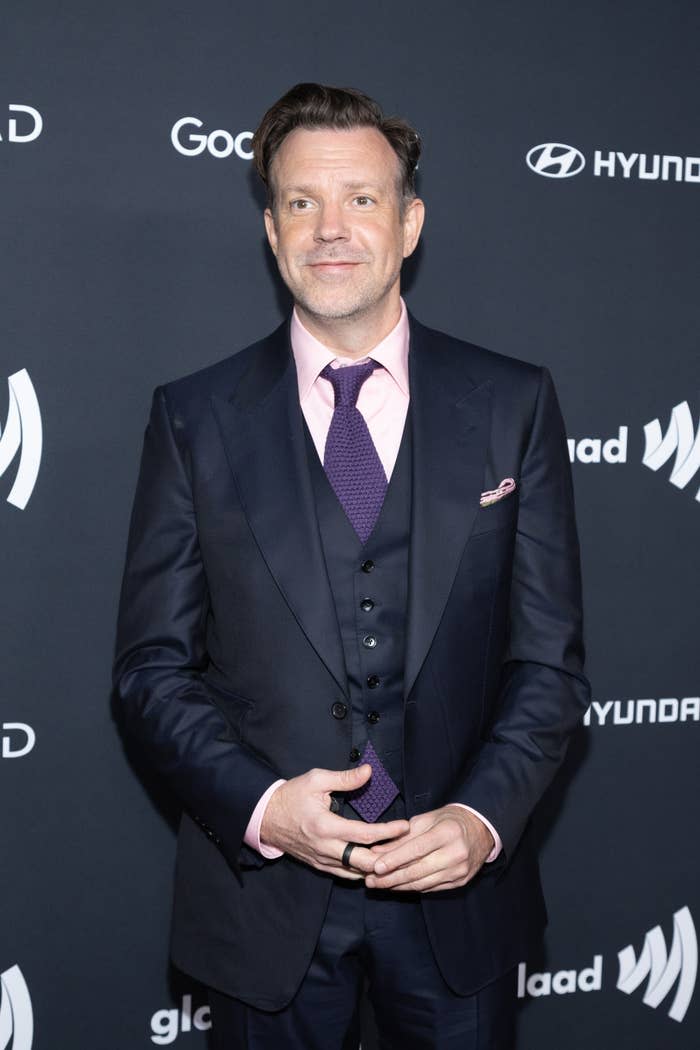 Jason Sudeikis on the red carpet wearing a dark three-piece suit with a light pink shirt and a dark tie at a GLAAD event