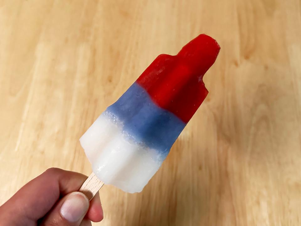 A hand holds a red, blue, and white ice pop in front of a wooden table