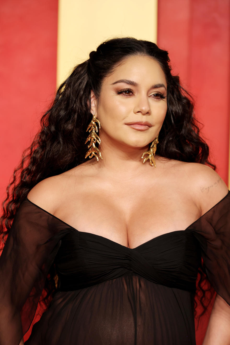 Vanessa Hudgens on the red carpet in a stylish black off-the-shoulder dress with sheer sleeves, wearing statement gold earrings