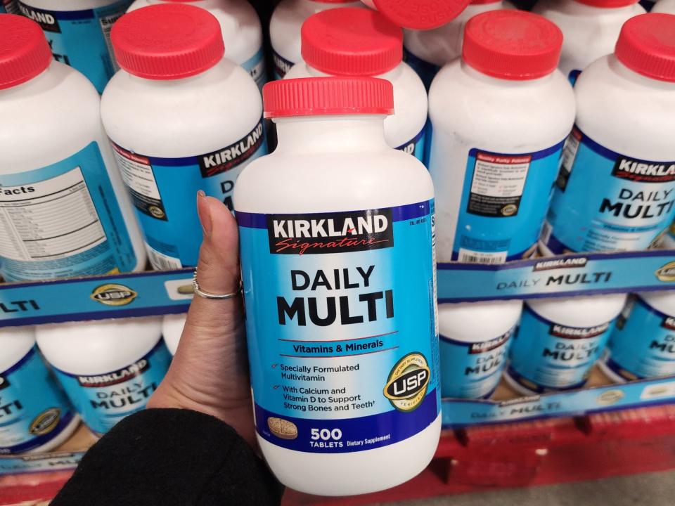 A hand holds a white bottle of multivitamins with a blue label and red cap