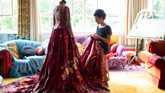 A dark haired woman is sat down, sewing on a long red dress in a living room