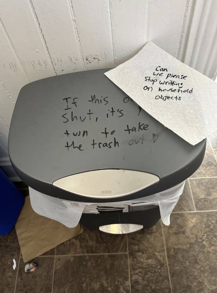 Handwritten note on a trash bin lid saying to shut it properly or take out the trash, with a "Can Please Stop Putting Sharpie on Household Objects" note