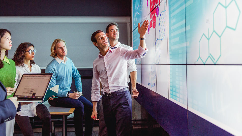 Group of business professionals in a dark room standing in front of a large data display screen with information.