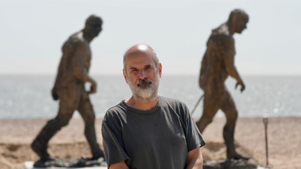 Laurence Edwards in a grey T-shirt standing in front of two statues