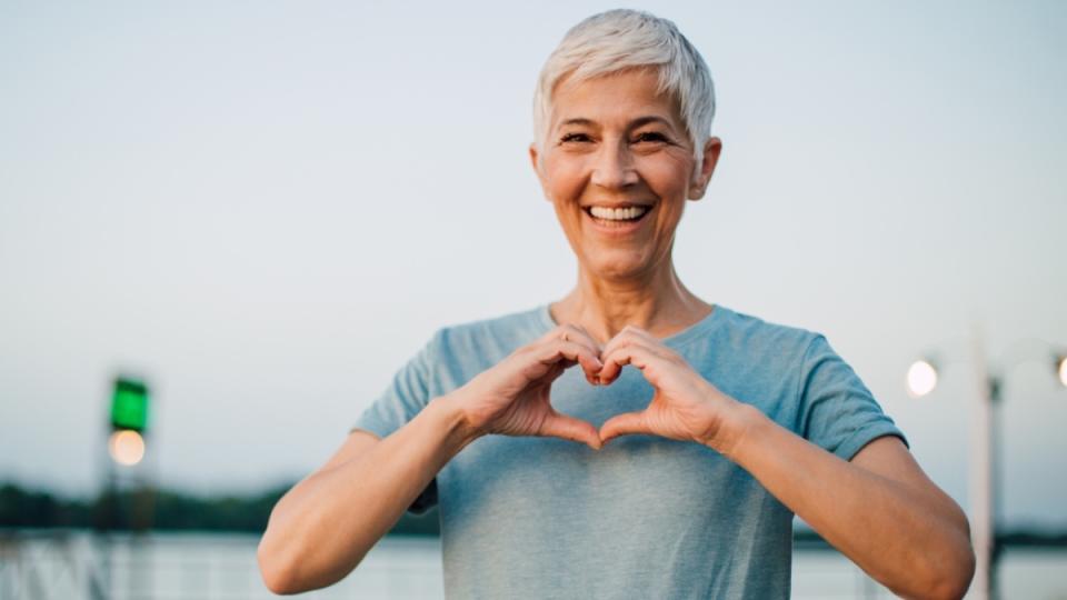 A woman with grey hair and a blue shirt making a heart shape with her hands