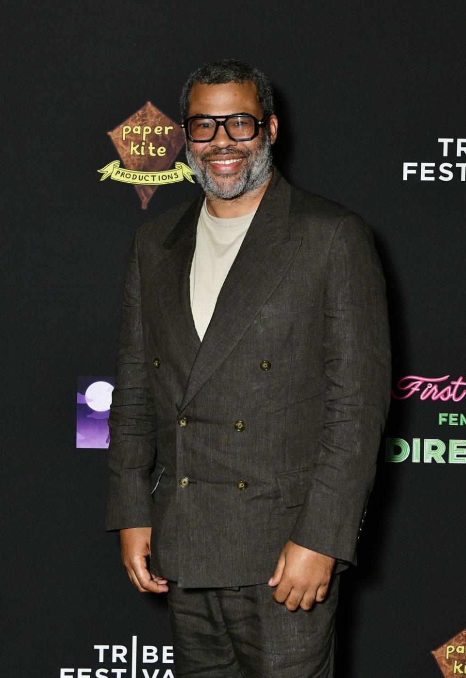 jordan peele, a man with black and grey hair and a black and grey beard, wearing black framed glasses, a tan shirt, and dark blazer while smiling for the camera