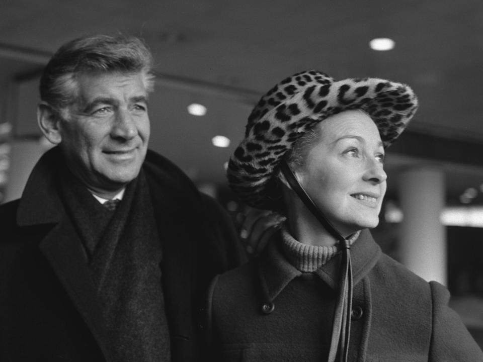 Leonard Bernstein, in a black coat and dark scarf, and Felicia Montealegre, in a peacoat and leopard print hat, smile and look at something off camera in this 1966 photo.