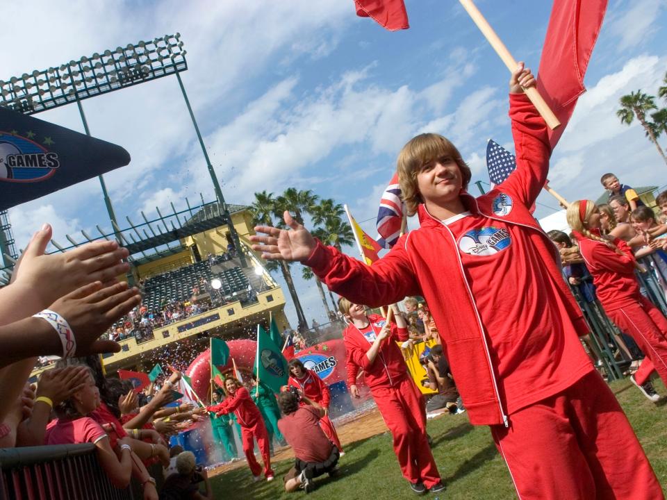 jason earles in red outfit with arm raised carrying flag in front of obstacle course at the disney channel games in 2007