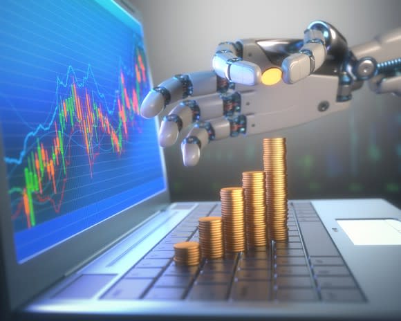 A robot hand stacking gold coins on the keyboard of a laptop, the screen of which is showing charts with high growth.