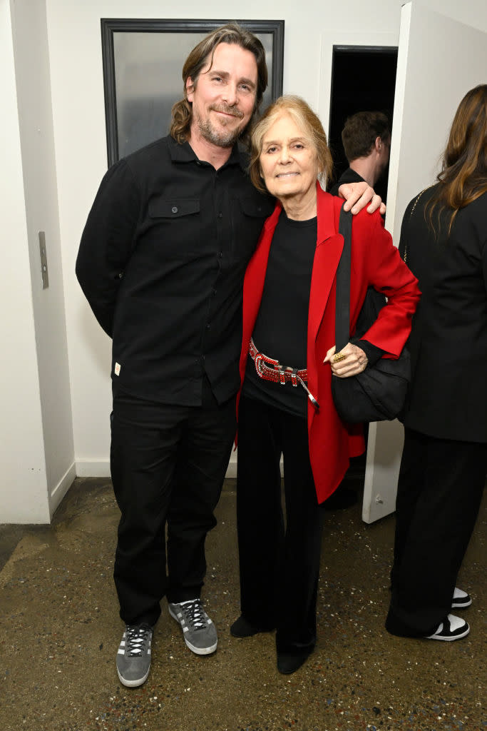 Two individuals posing for a photo; one in black attire with sneakers, the other in a red jacket and black pants, both smiling