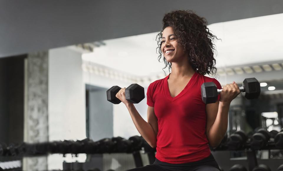 a woman with curly hair lifting dumbbells in a gym, wearing a red T-shirt