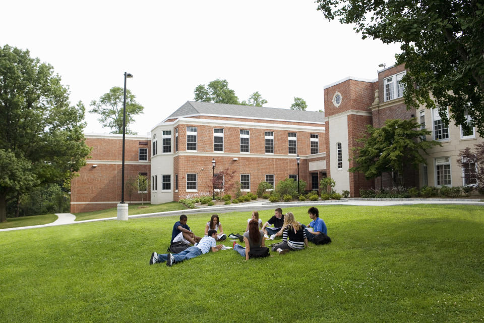 A group of people sitting on a grassy lawn in front of a large school building, engaged in conversation and appearing relaxed and happy