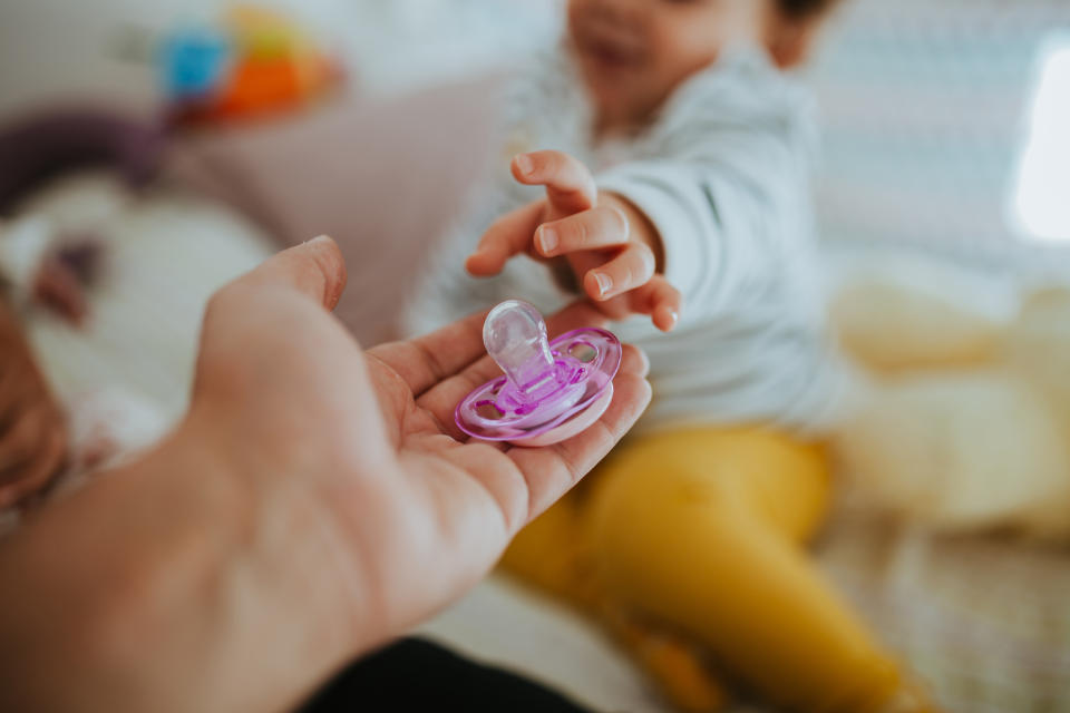 Adult hand holding a pacifier while a baby reaches for it in the background