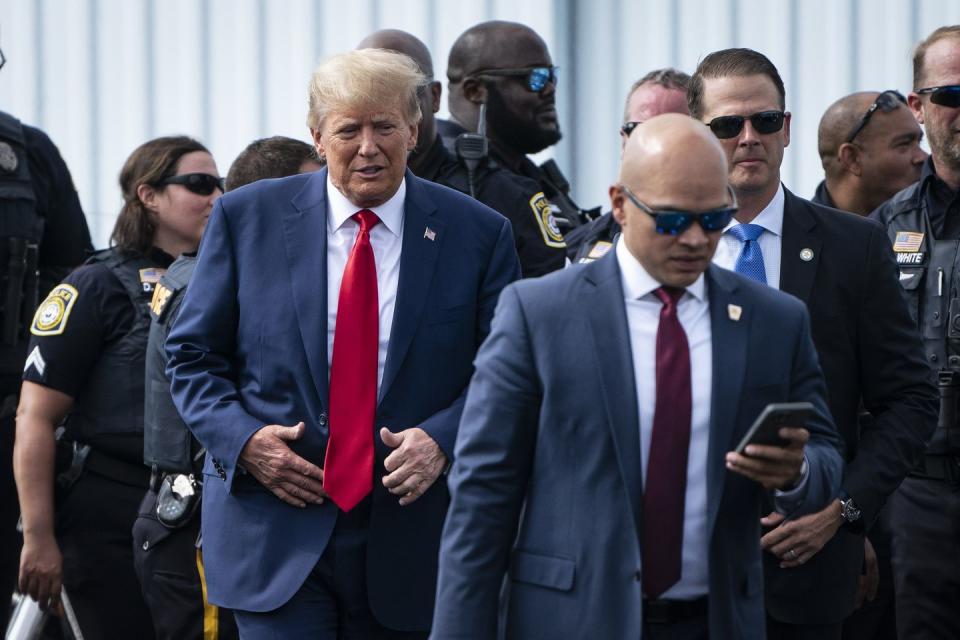 donald trump, wearing a blue suit and red tie, walks in front of several police officer along with walt nauta, who wears a blue suit and sunglasses while looking at a smartphone