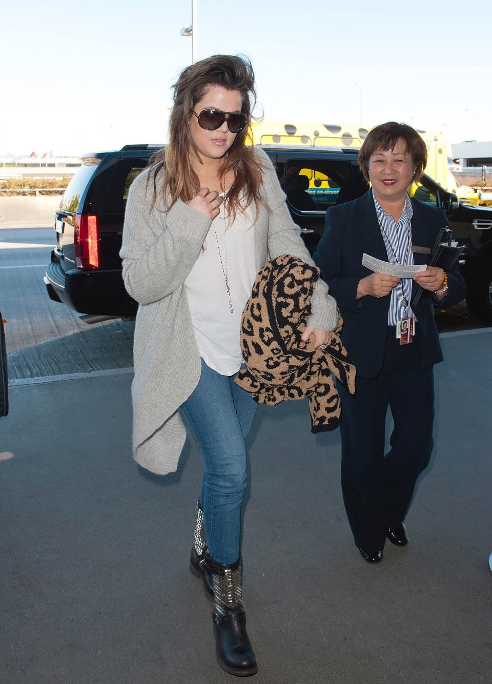 Khloé Kardashian at an airport, wearing jeans, a casual white top, a long cardigan, and black boots, holding a leopard print blanket, accompanied by an airport staff member