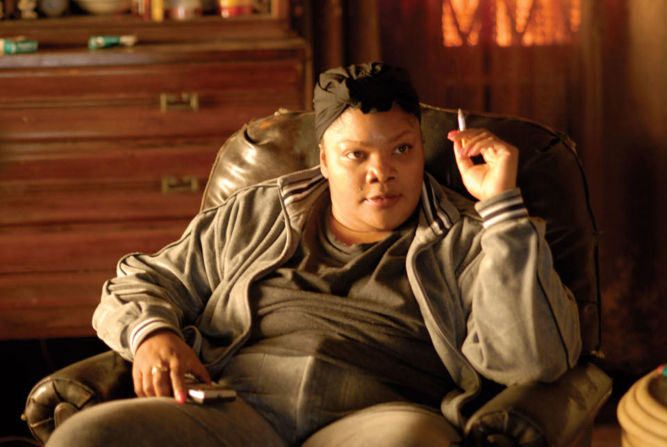 Mo'Nique in a casual outfit sits in a leather armchair, holding a cigarette in one hand and a TV remote in the other, appearing focused