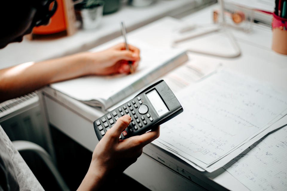 Person working at a desk, holding a scientific calculator in one hand and writing notes in a workbook with the other