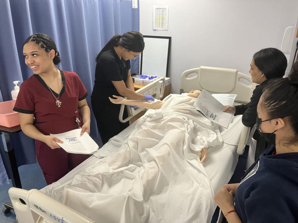 Diana Pimentel (left) listens to an advisor as RINI classmates (from left) Veronica Benitez, Joslin Lebron and Edilma Ramirez tend to a mock patient in a prep session for a certified nursing assistant exam. (Greg Toppo)<br><br>