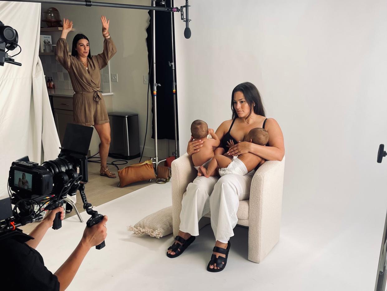 Ashley Graham breastfeeding her twins on set for her Bobbie video, sitting in a white chair wearing white pants and black sandals