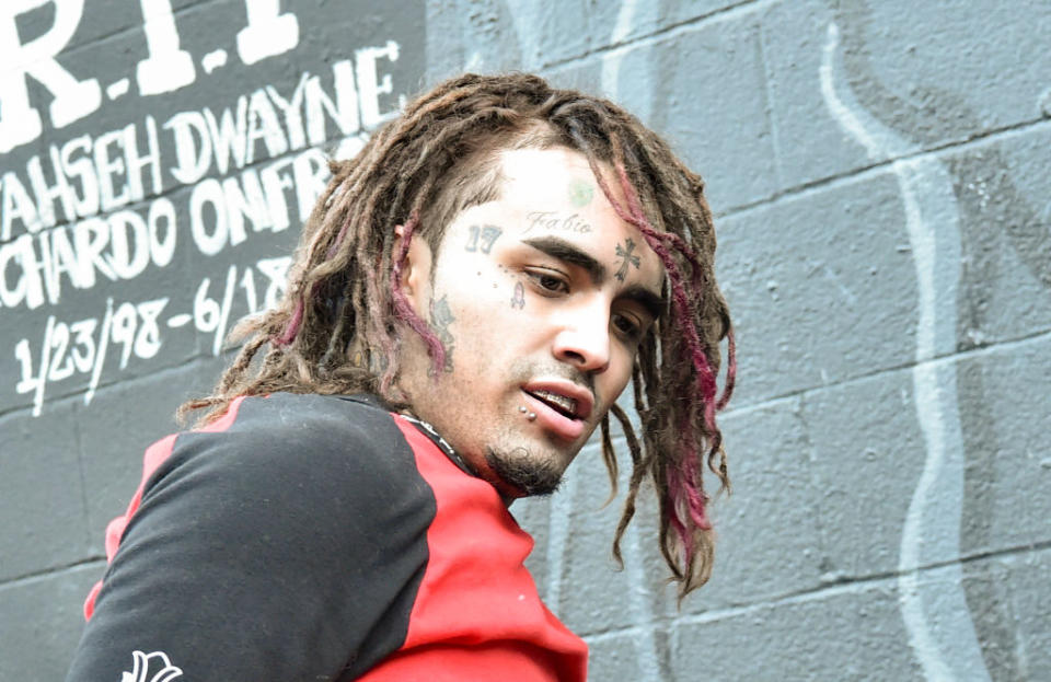 Lil Pump steht vor einer mit Graffiti bedeckten Wand und trägt ein rotes Hemd