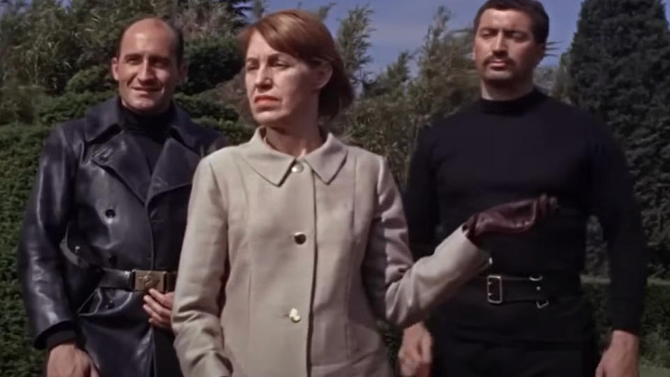 Lotte Lenya stands flanked by two Spectre agents on a sunny day in From Russia with Love.