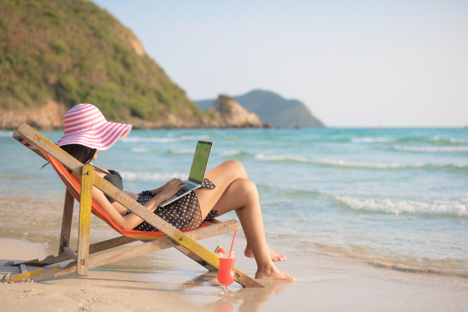 Young attractive woman in dress, sunglasses and hat, working in laptop on the beach