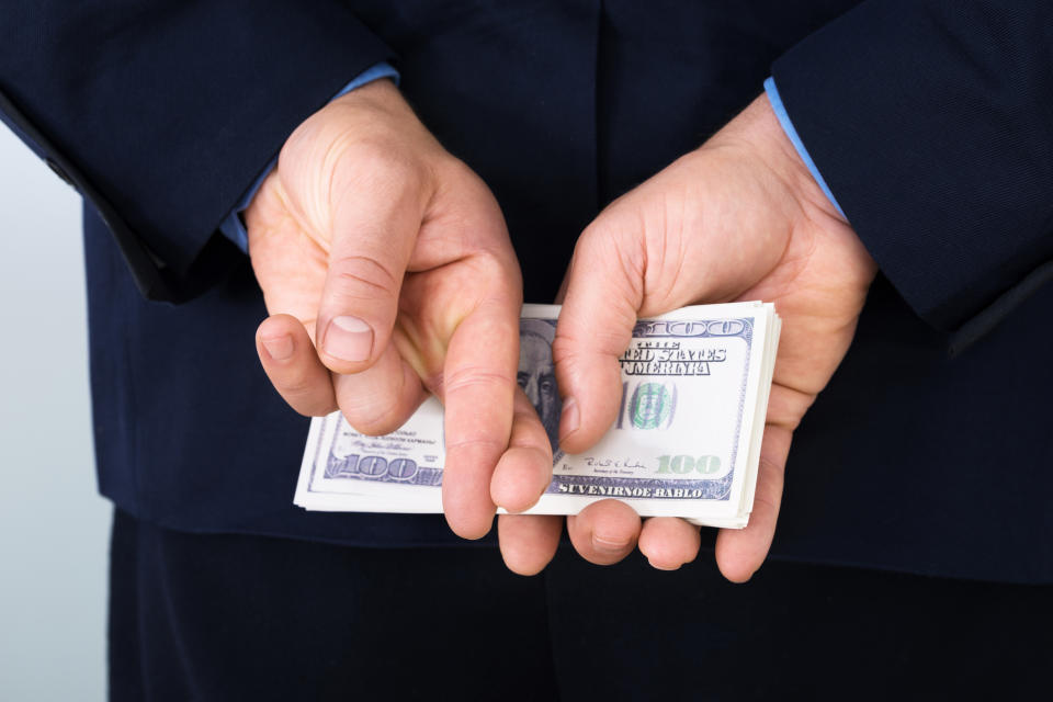 A man in a suit holding a stack of hundred-dollar bills behind his back, with his fingers crossed.