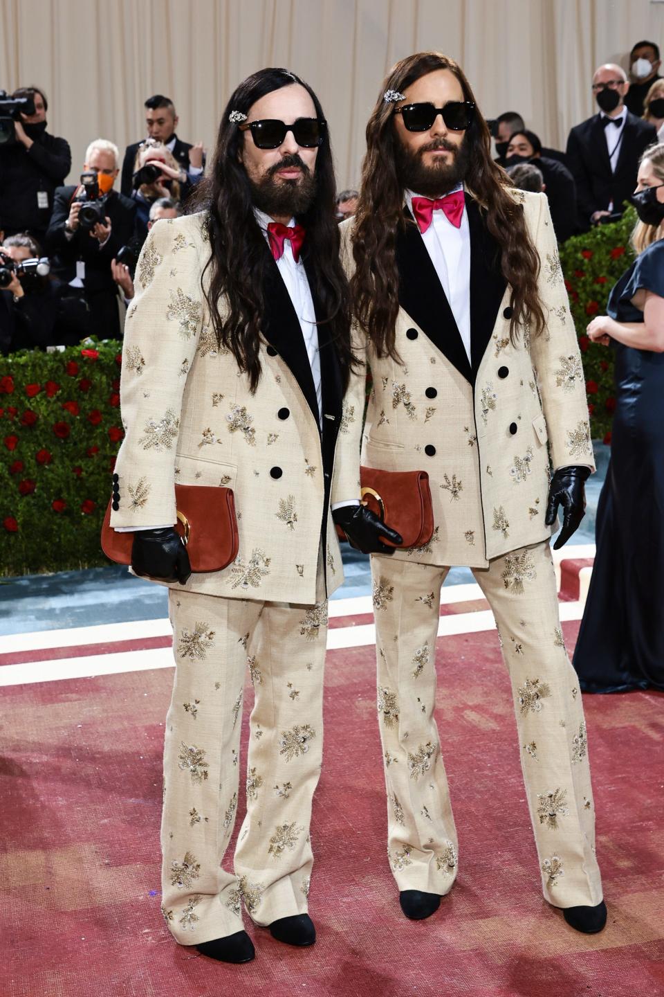 Alessandro and Jared Leto are wearing identical looks and are styled the same. They have long brown wavy hair with a beret, beards, and sunglasses on. They're wearing cream suits black lapels with pearl and bead embellishments throughout the jacket and pants. They have dark pink bowties, are holding leather clutch bags, have black leather gloves on, and are wearing pointed black shoes.