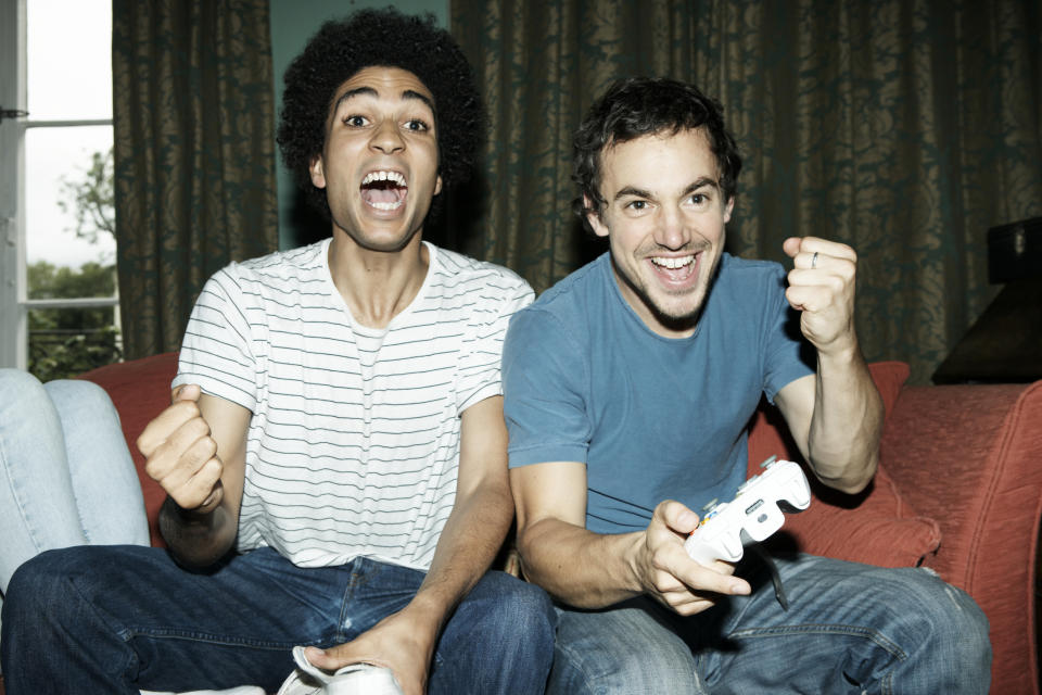 Two young men pump their fists in jubilation while sitting on a couch and holding game controllers.