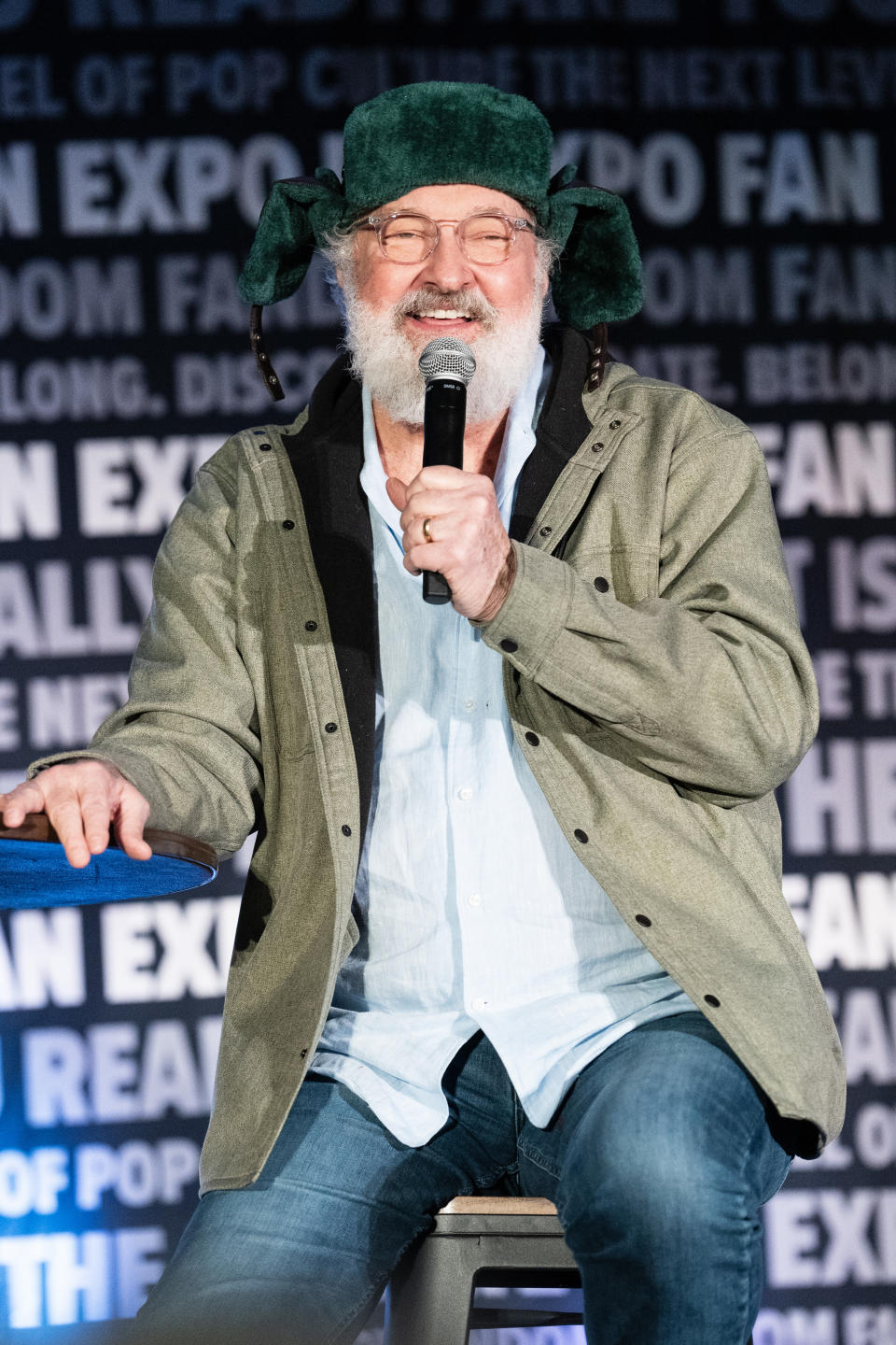 James Cromwell wearing a green hat with earmuffs and casual clothing speaks into a microphone at the Fan Expo event