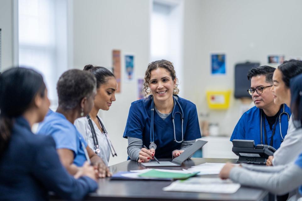 A small group of medical professionals gathers around a table.  They are all dressed professionally and there are files scattered around.