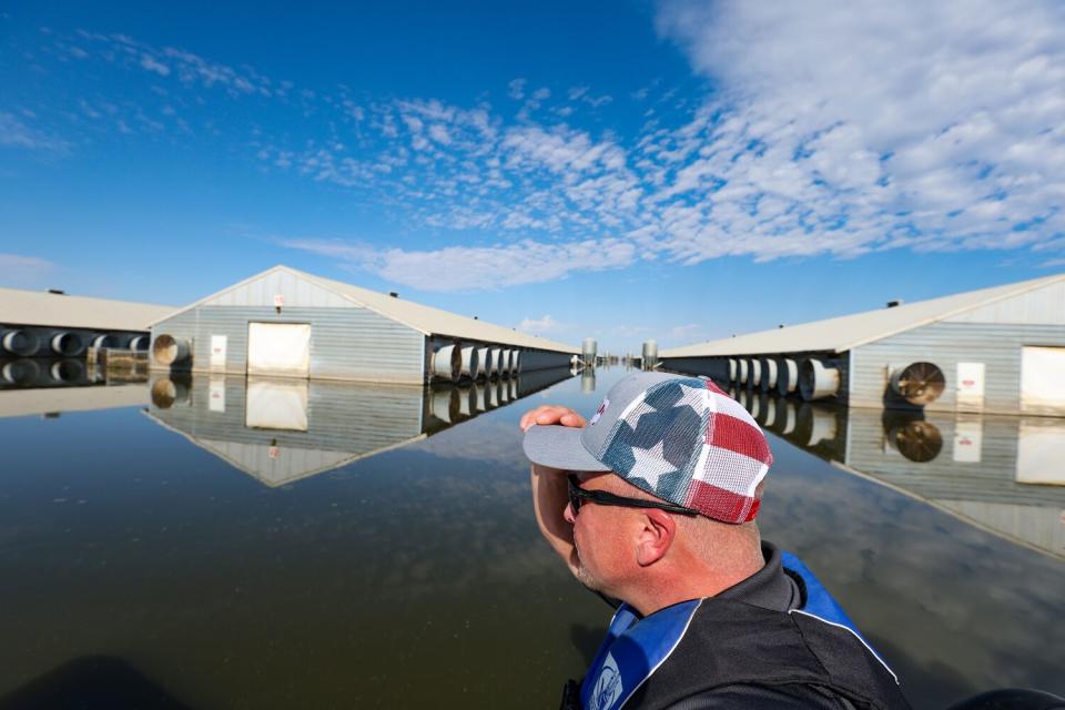 A man wearing a lifejacket and hat pilots an airboat past a flooded chicken ranch.