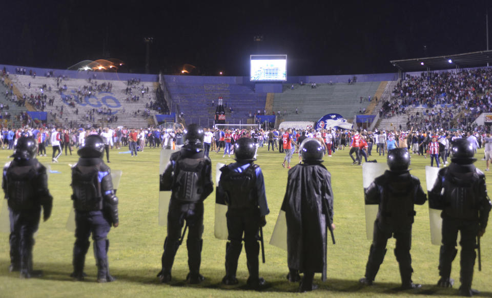 Police stand watch as soccer fans gather on the field to escape tear gas fired by police from inside the national stadium to the outside where deadly fights broke out between fans before the start of a game between Motagua and Olimpia in Tegucigalpa, Honduras, late Saturday, Aug. 17, 2019. The fight between fans of rival soccer teams outside the stadium left three people dead and led to the suspension of the game. (Victor Colindres/La Tribunal via AP)