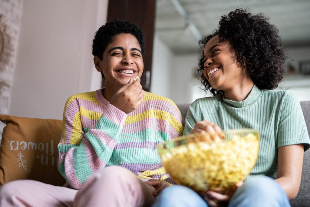 Two friends laugh as they watch television and eat popcorn together. 