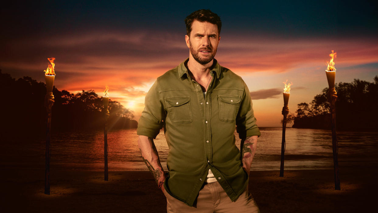  Joel Dommett photographed in front of lit torches on a beach at sundown for Survivor UK 2023. 