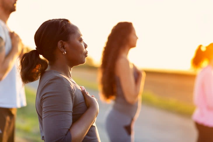 A group of people stands with their hands on their hearts facing the sunset. A black woman with a ponytail wearing a gray shirt is in the foreground.