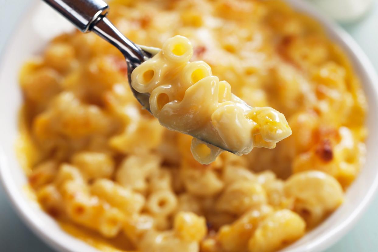 Closeup of crock-pot mac & cheese on a fork over a white porcelain bowl of it, selective focus on a white table