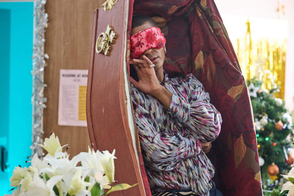 Stephen (Layton Williams) lies upright inside a luxuriously-upholstered coffin, positioned vertically. He is wearing a floral eye mask.