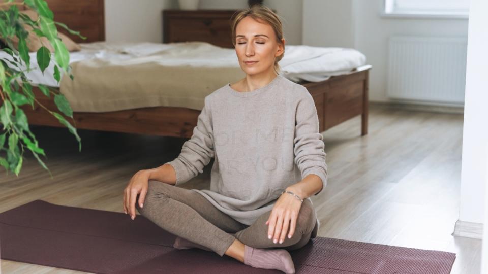 A woman sitting on a yoga mat with closed eyes in her bedroom as she practices a work out in the evening time