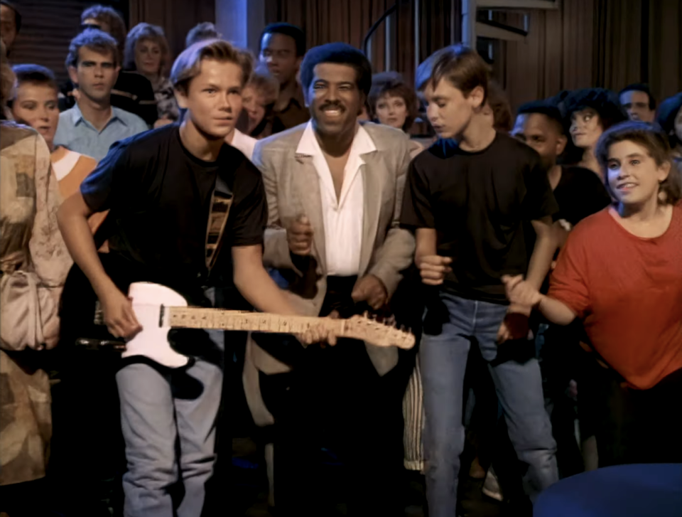 a teenager playing guitar while a crowd surrounds him